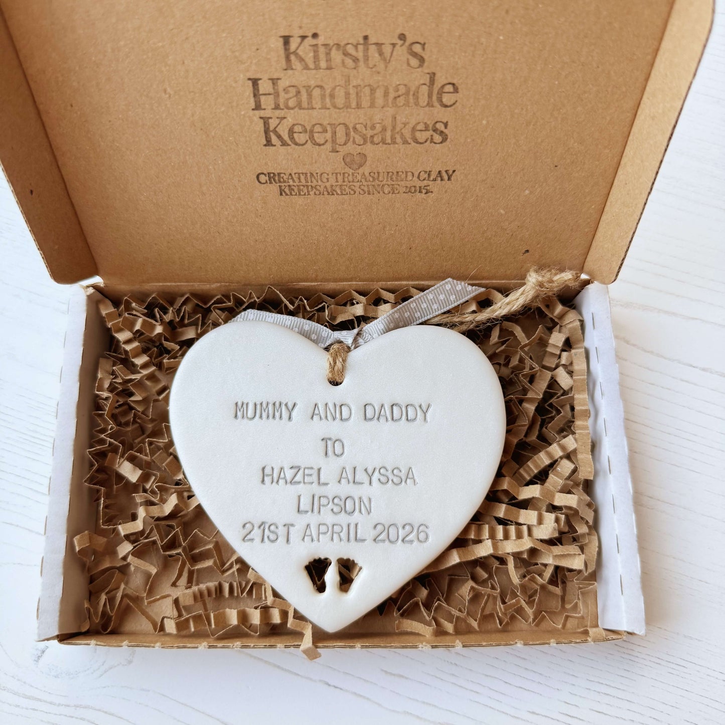 Pearlised white clay hanging heart with baby feet cut out of the bottom and grey personalisation, the heart is personalised with MUMMY & DADDY TO HAZEL ALYSSA LIPSON 21ST APRIL 2026