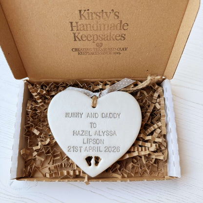 Pearlised white clay hanging heart with baby feet cut out of the bottom and grey personalisation, the heart is personalised with MUMMY & DADDY TO HAZEL ALYSSA LIPSON 21ST APRIL 2026