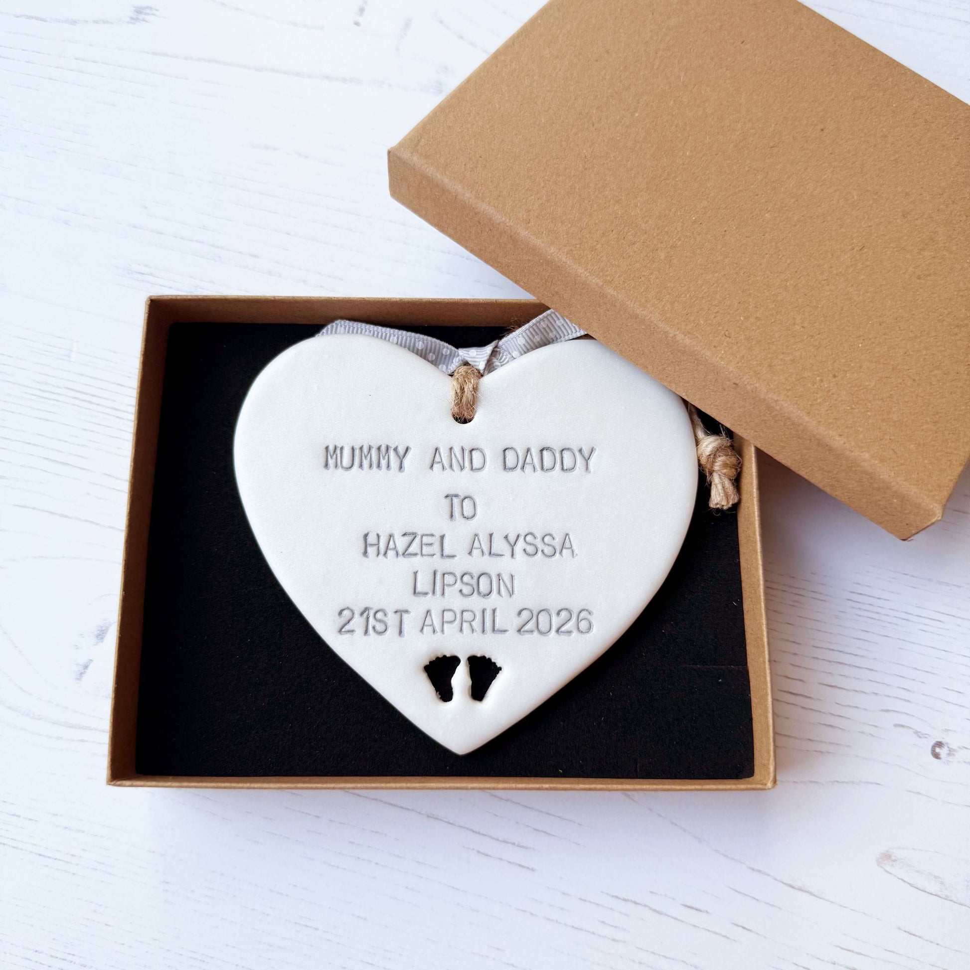 Pearlised white clay hanging heart with baby feet cut out of the bottom and grey personalisation, the heart is personalised with MUMMY & DADDY TO HAZEL ALYSSA LIPSON 21ST APRIL 2026