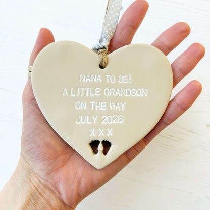 Beige clay hanging heart with baby feet cut out of the bottom and white personalisation, the heart is personalised with NANA TO BE! A LITTLE GRANDSON ON THE WAY JULY 2026 XXX