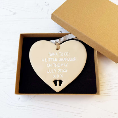 Beige clay hanging heart with baby feet cut out of the bottom and white personalisation, the heart is personalised with NANA TO BE! A LITTLE GRANDSON ON THE WAY JULY 2026 XXX