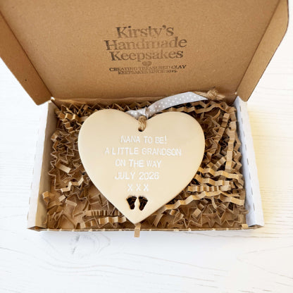 Beige clay hanging heart with baby feet cut out of the bottom and white personalisation, the heart is personalised with NANA TO BE! A LITTLE GRANDSON ON THE WAY JULY 2026 XXX