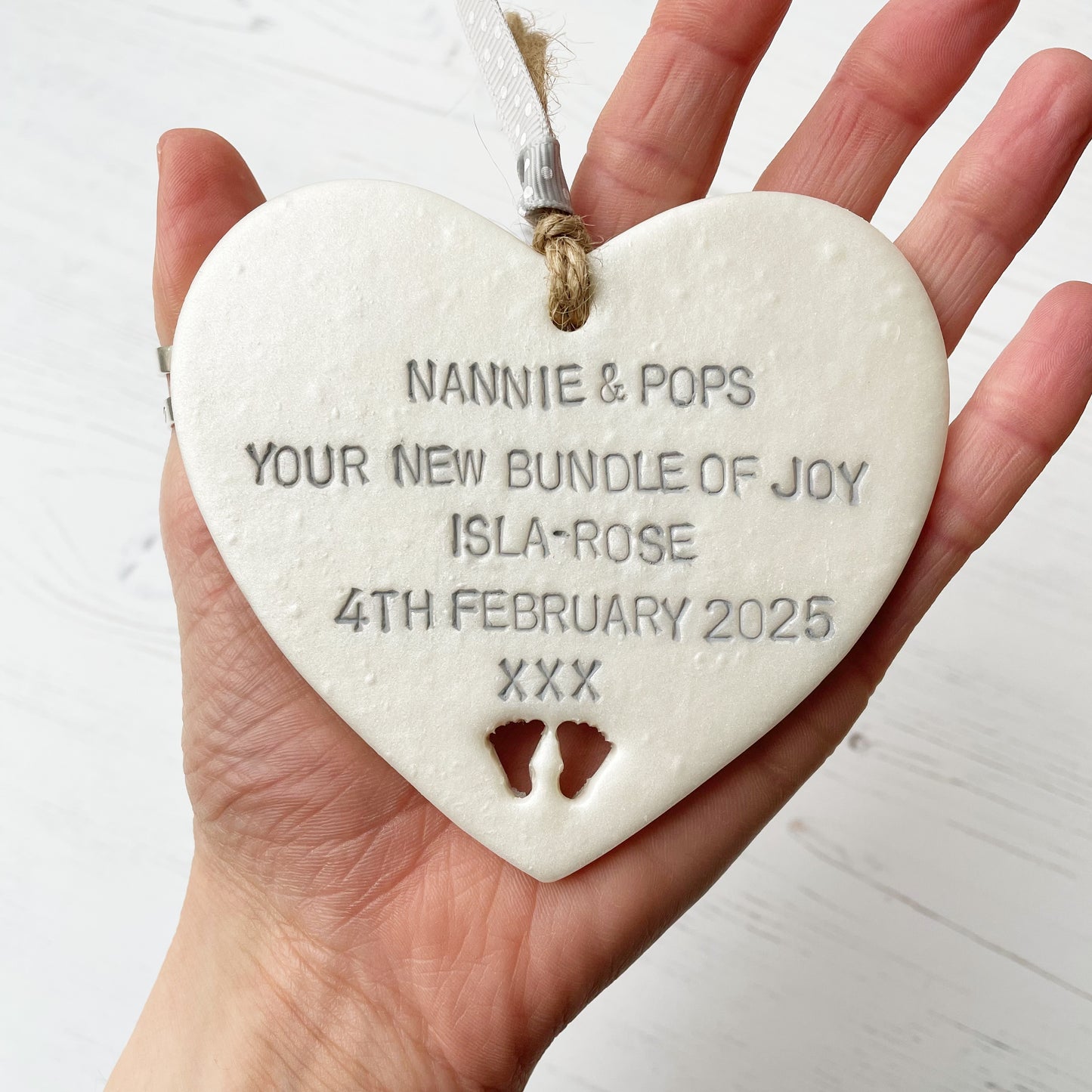 Pearlised white clay hanging heart with baby feet cut out of the bottom and grey personalisation, the heart is personalised with NANNIE & POPS YOUR NEW BUNDLE OF JOY ISLA-ROSE 4TH FEBRUARY 2025 XXX