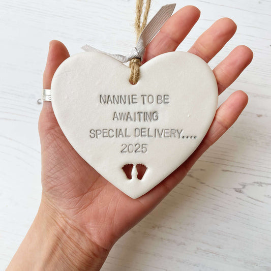 Pearlised white clay hanging heart with baby feet cut out of the bottom and grey personalisation, the heart is personalised with NANNIE TO BE AWAITING SOECIAL DELIVERY…. 2025