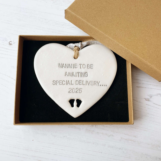 Pearlised white clay hanging heart with baby feet cut out of the bottom and grey personalisation, the heart is personalised with NANNIE TO BE AWAITING SOECIAL DELIVERY…. 2025