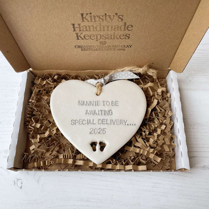 Pearlised white clay hanging heart with baby feet cut out of the bottom and grey personalisation, the heart is personalised with NANNIE TO BE AWAITING SOECIAL DELIVERY…. 2025