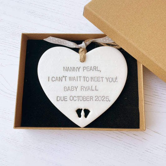 Pearlised white clay hanging heart with baby feet cut out of the bottom and grey personalisation, the heart is personalised with NANNY PEARL, I CAN’T WAIT TO MEET YOU! BABY RYALL DUE OCTOBER 2025