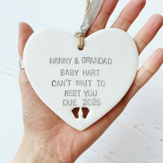 Pearlised white clay hanging heart with baby feet cut out of the bottom and grey personalisation, the heart is personalised with NANNY & GRANDAD BABY HART CAN’T WAIT TO MEET YOU DUE 2025