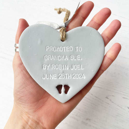 Grey clay hanging heart with baby feet cut out of the bottom and white personalisation, the heart is personalised with PROMOTED TO GRANDMA SUE. BY ROBIN JOEL JUNE ON 25TH 2024