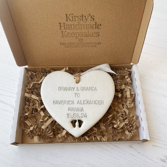 Pearlised white clay hanging heart with baby feet cut out of the bottom and grey personalisation, the heart is personalised with GRANNY & GRANDA TO MAVERICK ALEXANDER HANNA 15.08.24