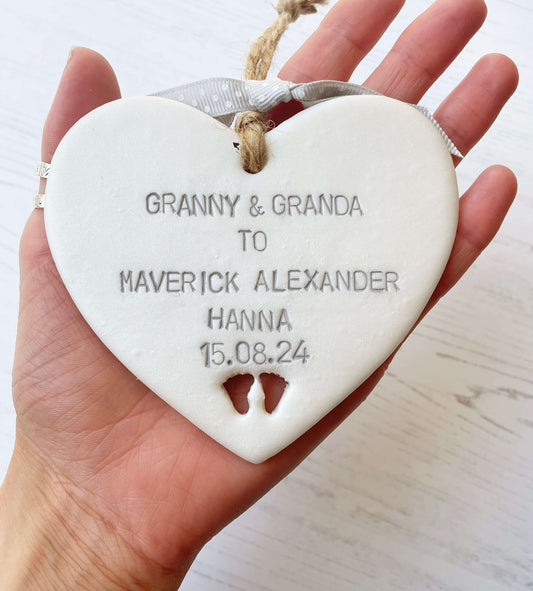 Pearlised white clay hanging heart with baby feet cut out of the bottom and grey personalisation, the heart is personalised with GRANNY & GRANDA TO MAVERICK ALEXANDER HANNA 15.08.24