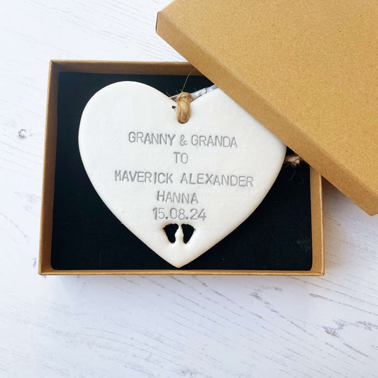 Pearlised white clay hanging heart with baby feet cut out of the bottom and grey personalisation, the heart is personalised with GRANNY & GRANDA TO MAVERICK ALEXANDER HANNA 15.08.24
