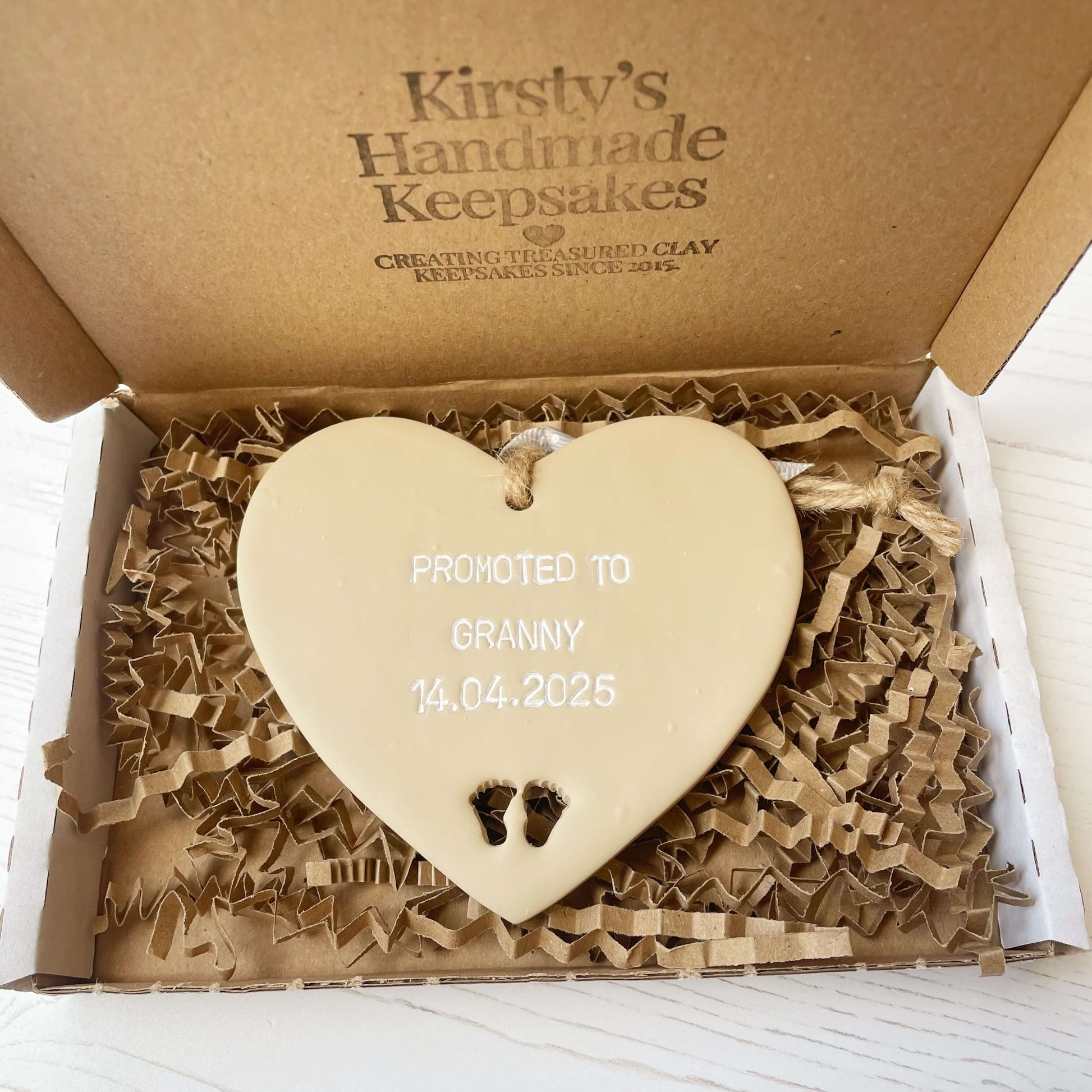 Beige clay hanging heart with baby feet cut out of the bottom and white personalisation, the heart is personalised with PROMOTED TO GRANNY 14.04.2025