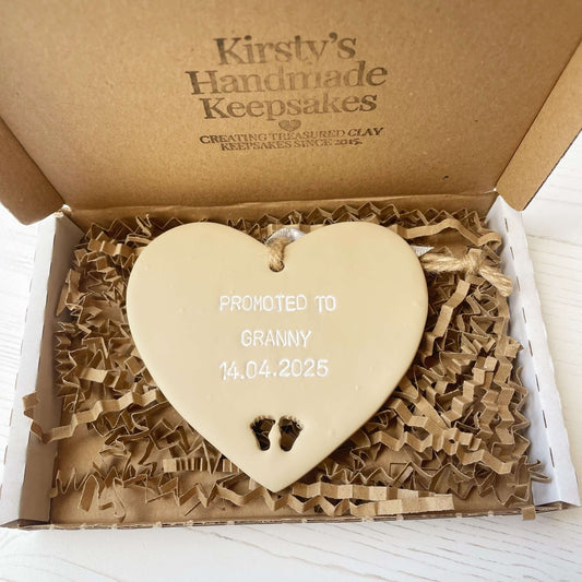 Beige clay hanging heart with baby feet cut out of the bottom and white personalisation, the heart is personalised with PROMOTED TO GRANNY 14.04.2025