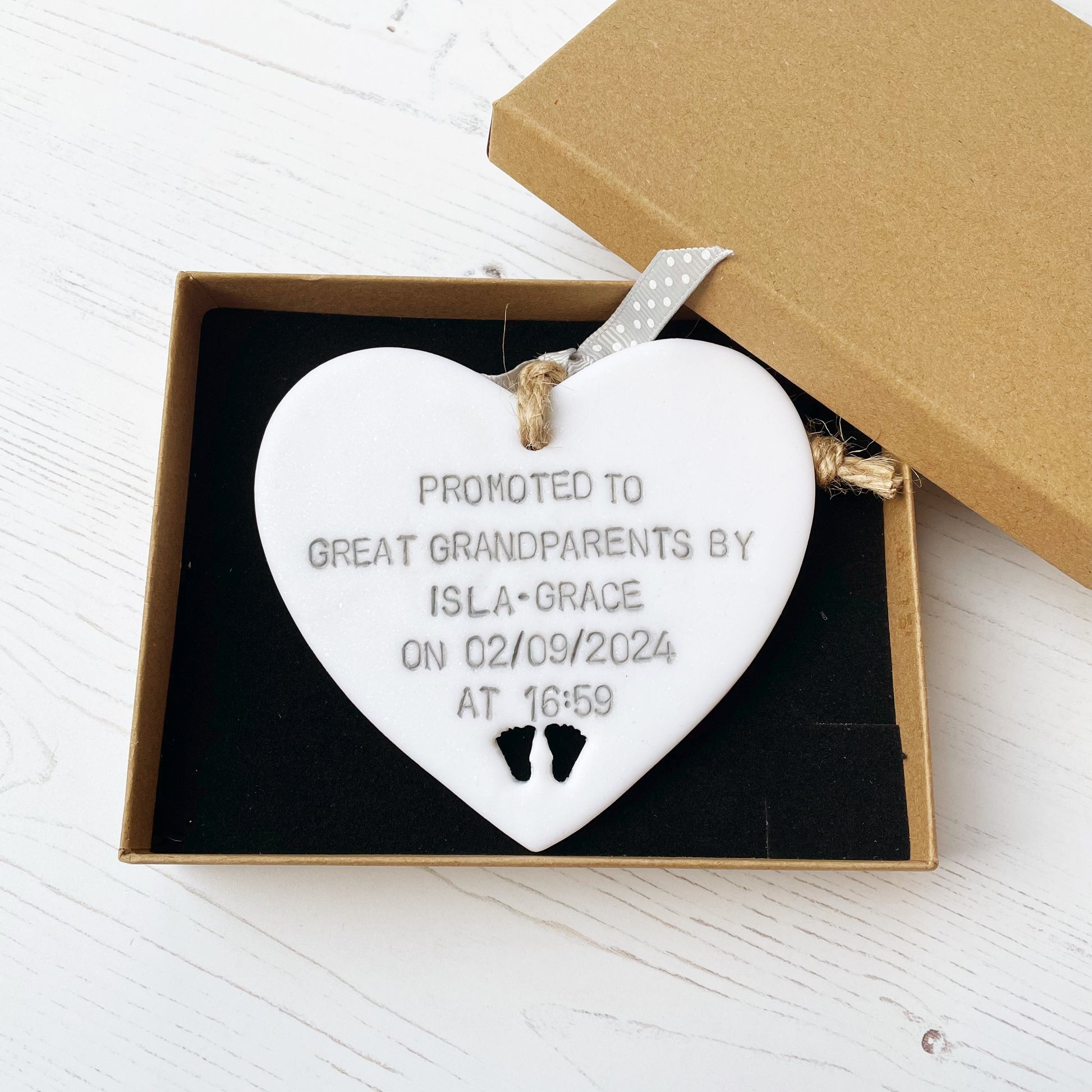 Pearlised white clay hanging heart with baby feet cut out of the bottom and grey personalisation, the heart is personalised with PROMOTED TO GREAT GRANDPARENTS BY ISLA-GRACE ON 02/09/2024 AT 16:59