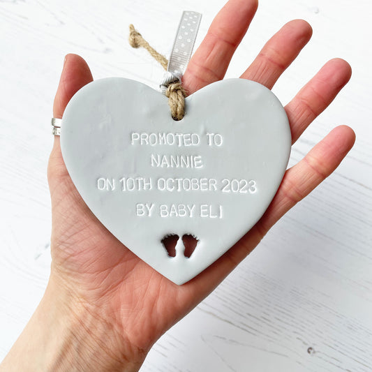 Grey clay hanging heart with baby feet cut out of the bottom and white personalisation, the heart is personalised with PROMOTED TO NANNIE ON 10TH OCTOBER 2023 BY BABY ELI