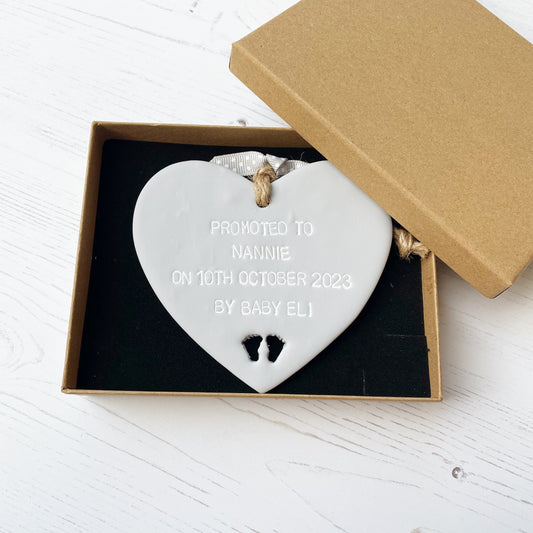 Grey clay hanging heart with baby feet cut out of the bottom and white personalisation, the heart is personalised with PROMOTED TO NANNIE ON 10TH OCTOBER 2023 BY BABY ELI