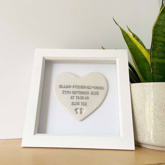Personalised baby keepsake gift, pearlised white clay heart with baby feet cut out at the bottom of the heart and grey personalisation in a white box frame, the heart is personalised with the baby’s name, date of birth, weight and time