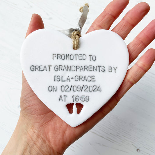 Pearlised white clay hanging heart with baby feet cut out of the bottom and grey personalisation, the heart is personalised with PROMOTED TO GREAT GRANDPARENTS BY ISLA-GRACE ON 02/09/2024 AT 16:59