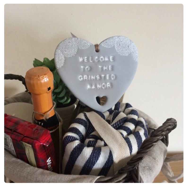 A grey polymer clay heart reading 'WELCOME TO THE GRINSTED MANOR with a small heart cutout, displayed in a woven basket with a striped cloth and other home items against a plain wall.