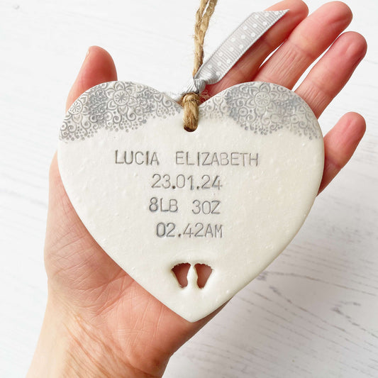 Personalised baby keepsake gift, pearlised white clay hanging heart with baby feet cut out at the bottom of the heart and a grey lace edge at the top of the heart, the heart is personalised with the baby’s name, date of birth, weight and time