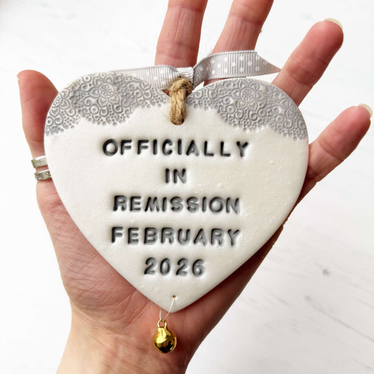 Personalised cancer survivor gift, pearlised white clay hanging heart with a grey lace edge at the top of the heart and a gold bell hanging below, the heart is personalised with OFFICIALLY IN REMISSION FEBRUARY 2026