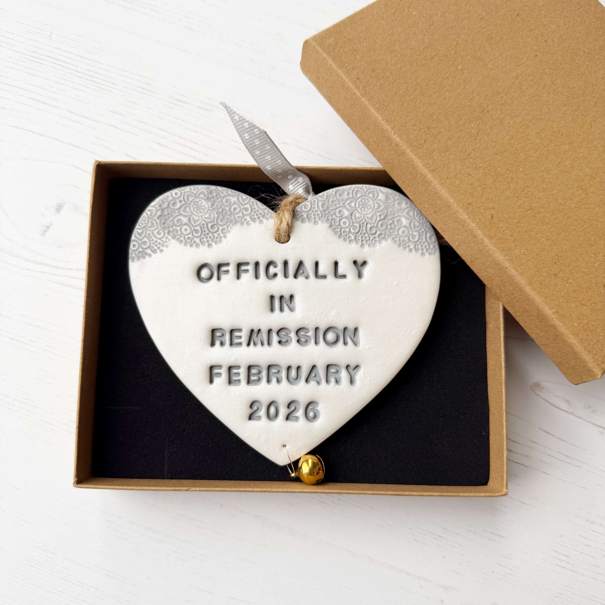 Personalised cancer survivor gift, pearlised white clay hanging heart with a grey lace edge at the top of the heart and a gold bell hanging below, the heart is personalised with OFFICIALLY IN REMISSION FEBRUARY 2026