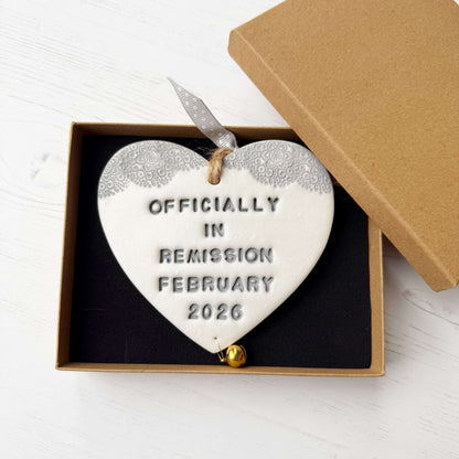 Personalised cancer survivor gift, pearlised white clay hanging heart with a grey lace edge at the top of the heart and a gold bell hanging below, the heart is personalised with OFFICIALLY IN REMISSION FEBRUARY 2026