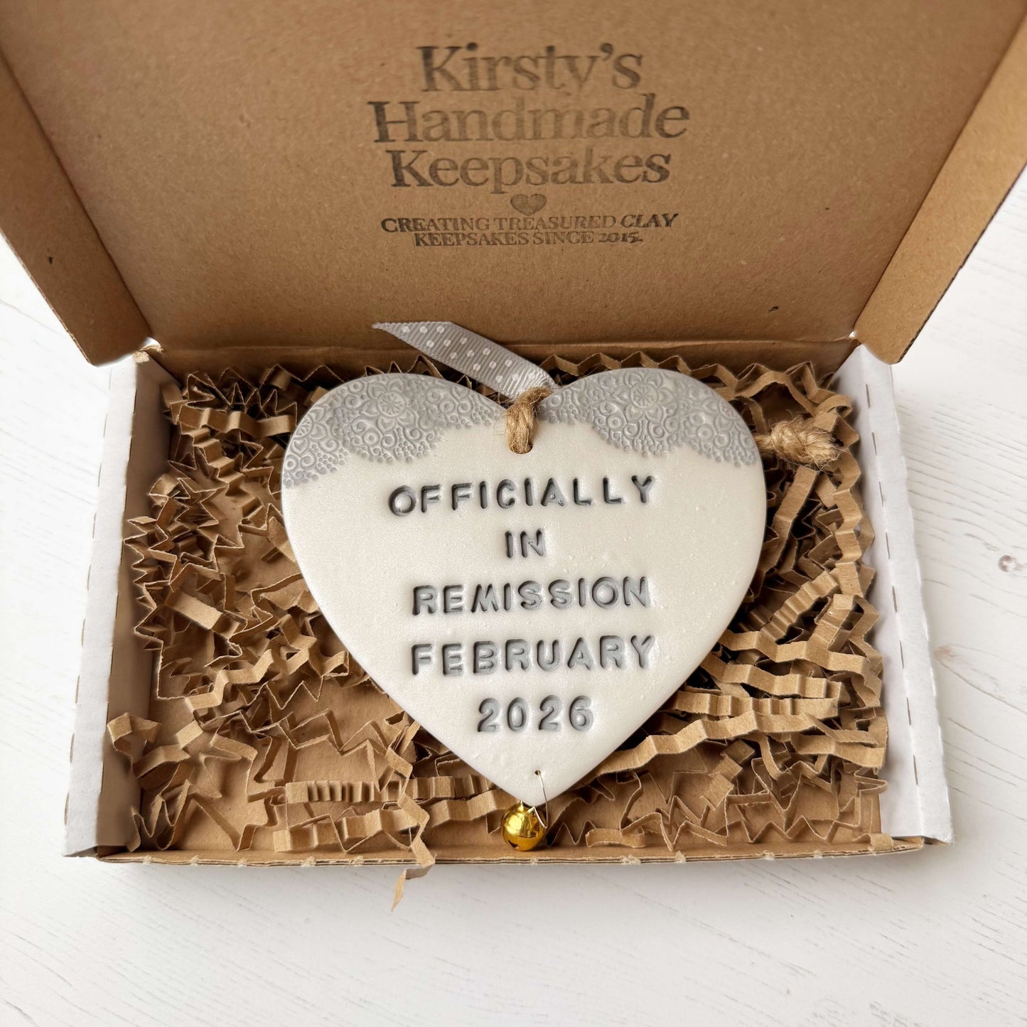 Personalised cancer survivor gift, pearlised white clay hanging heart with a grey lace edge at the top of the heart and a gold bell hanging below, the heart is personalised with OFFICIALLY IN REMISSION FEBRUARY 2026