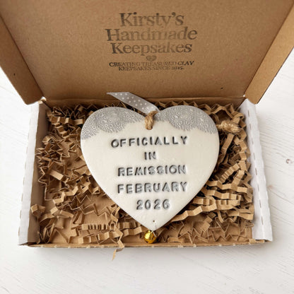 Personalised cancer survivor gift, pearlised white clay hanging heart with a grey lace edge at the top of the heart and a gold bell hanging below, the heart is personalised with OFFICIALLY IN REMISSION FEBRUARY 2026