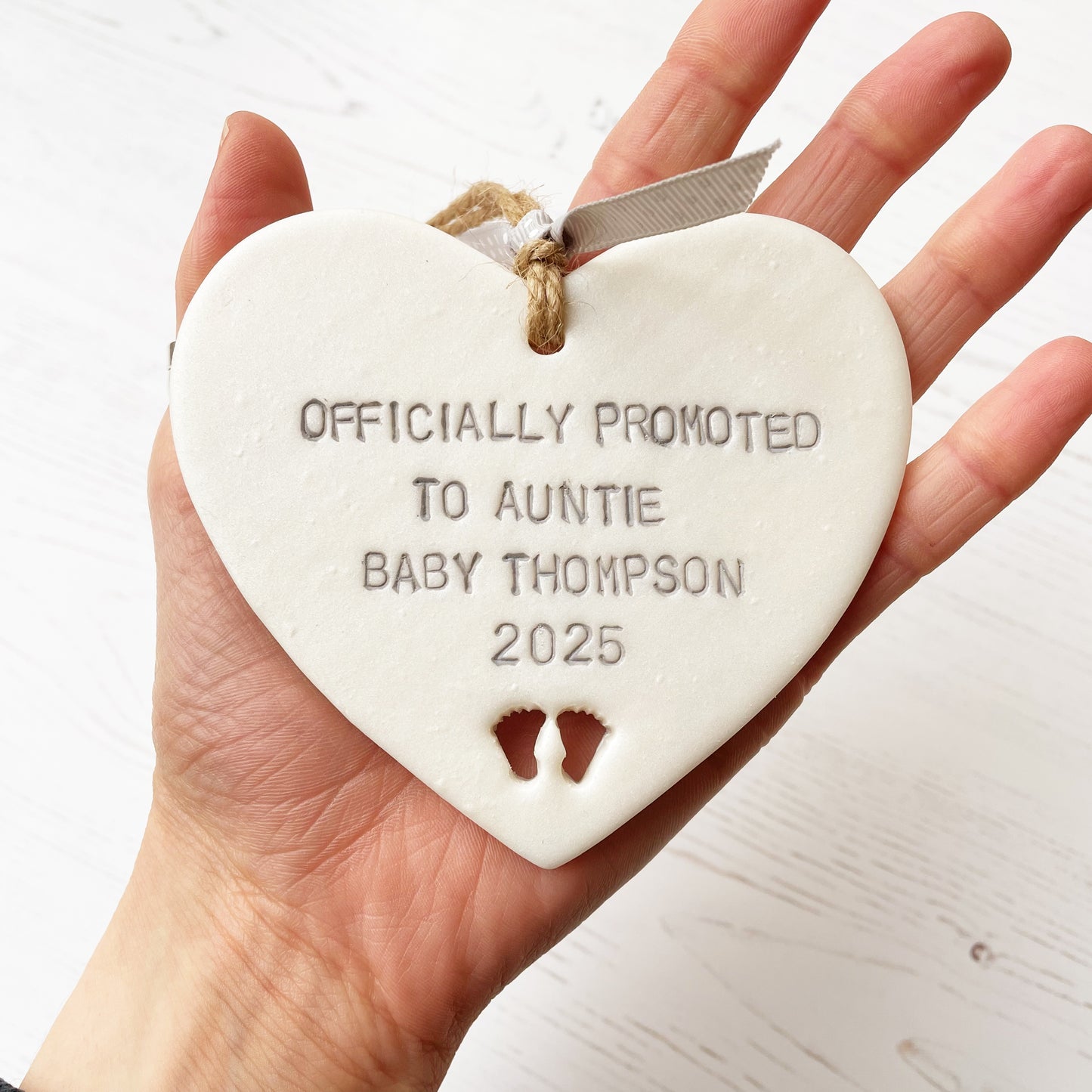 Pearlised white clay hanging heart with baby feet cut out of the bottom and grey personalisation, the heart is personalised with OFFICIALLY PROMOTED TO AUNTIE BABY THOMPSON 2025