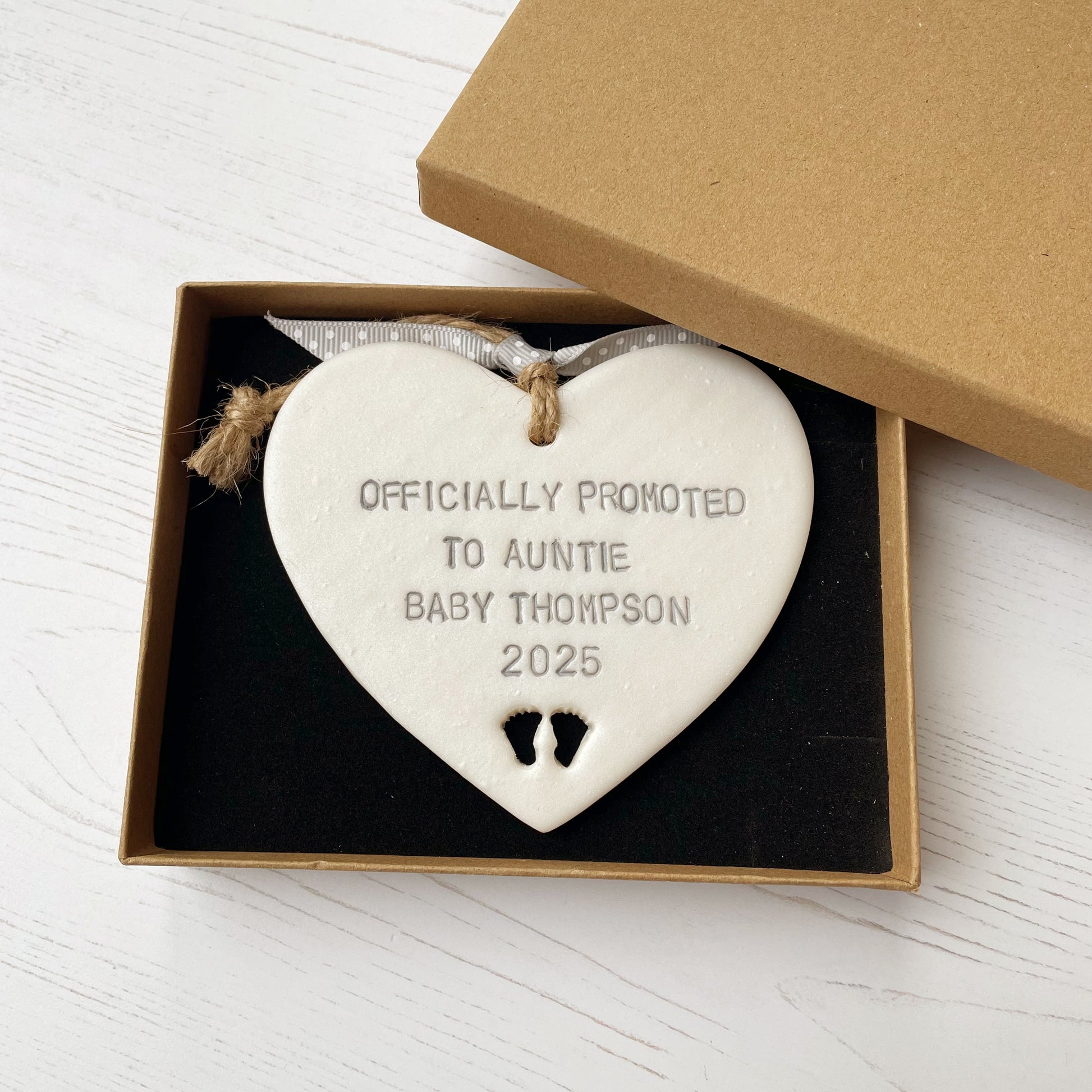 Pearlised white clay hanging heart with baby feet cut out of the bottom and grey personalisation, the heart is personalised with OFFICIALLY PROMOTED TO AUNTIE BABY THOMPSON 2025