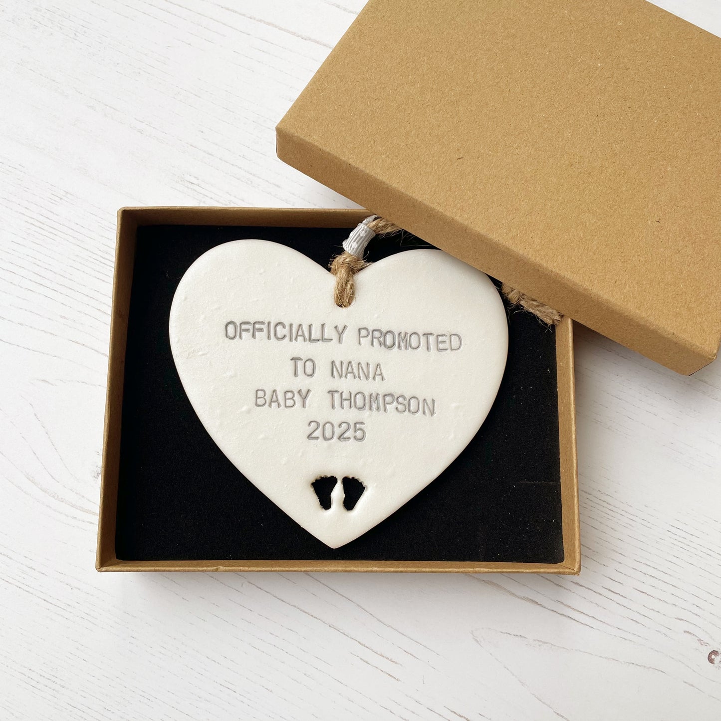 Pearlised white clay hanging heart with baby feet cut out of the bottom and grey personalisation, the heart is personalised with OFFICIALLY PROMOTED TO NANA BABY THOMPSON 2025