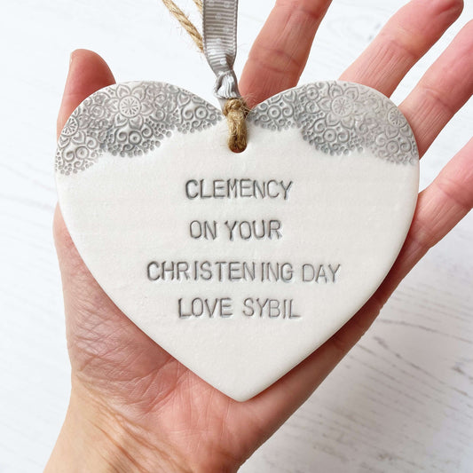 Personalised Christening baptism gift, Pearlised white clay hanging heart with a grey lace edge at the top of the heart, the heart is personalised with CLEMENCY ON YOUR CHRISTENING DAY LOVE SYBIL