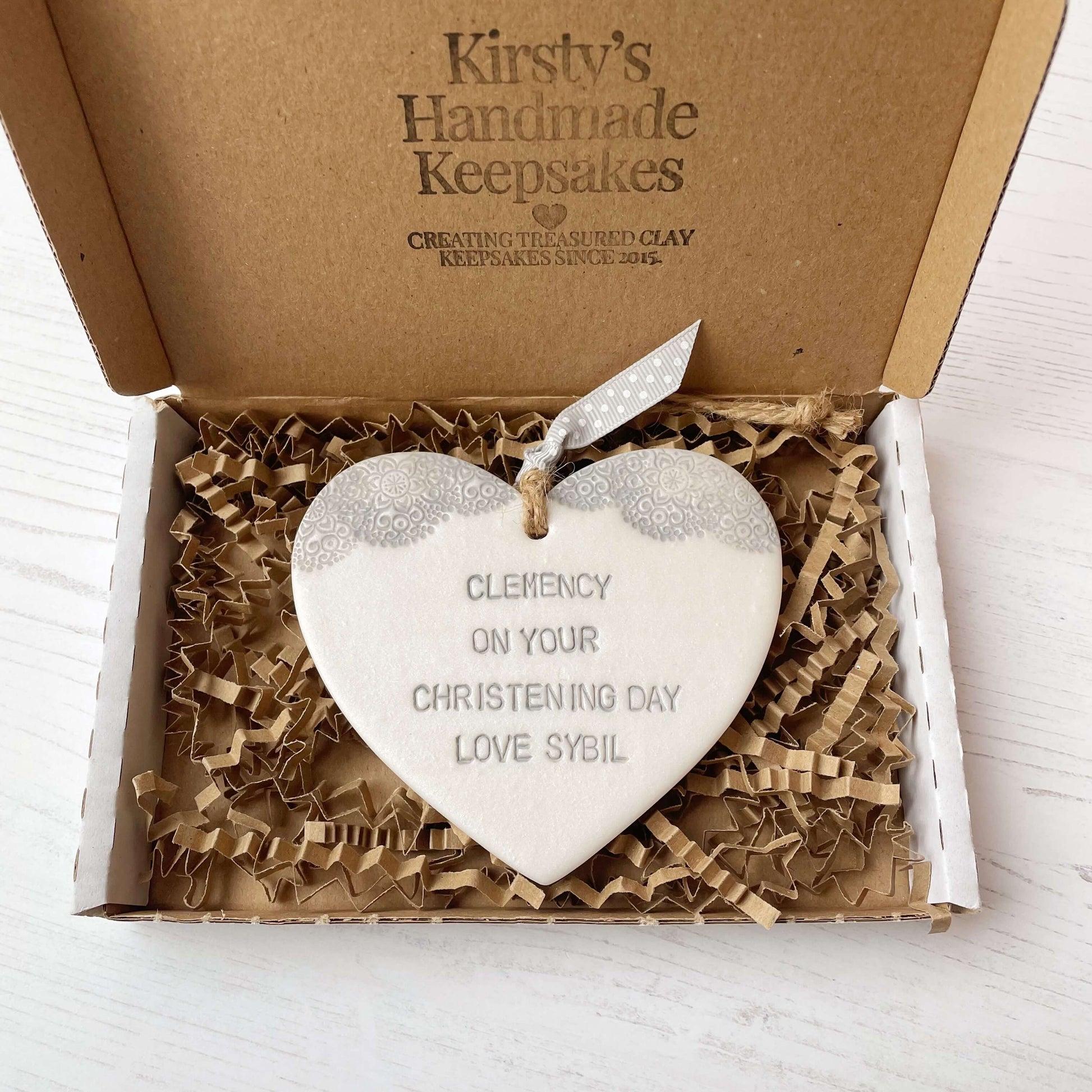 Personalised Christening baptism gift, Pearlised white clay hanging heart with a grey lace edge at the top of the heart, the heart is personalised with CLEMENCY ON YOUR CHRISTENING DAY LOVE SYBIL