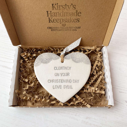 Personalised Christening baptism gift, Pearlised white clay hanging heart with a grey lace edge at the top of the heart, the heart is personalised with CLEMENCY ON YOUR CHRISTENING DAY LOVE SYBIL