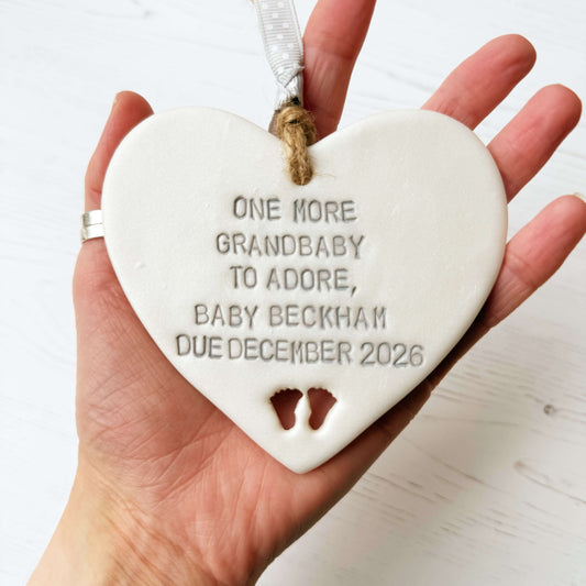 Pearlised white clay hanging heart with baby feet cut out of the bottom and grey personalisation, the heart is personalised with ONE MORE GRANDBABY TO ADORE BABY BECKHAM DUE DECEMBER 2026