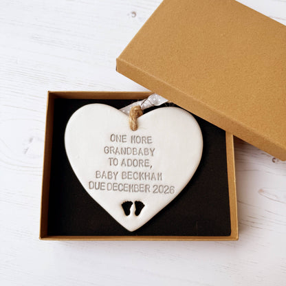 Pearlised white clay hanging heart with baby feet cut out of the bottom and grey personalisation, the heart is personalised with ONE MORE GRANDBABY TO ADORE BABY BECKHAM DUE DECEMBER 2026