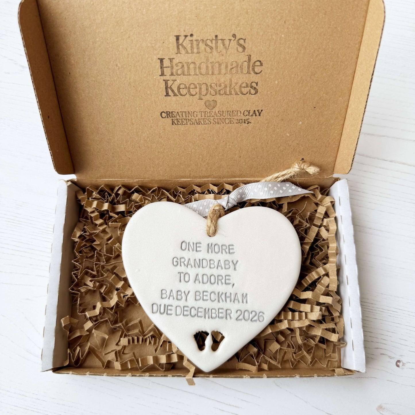 Pearlised white clay hanging heart with baby feet cut out of the bottom and grey personalisation, the heart is personalised with ONE MORE GRANDBABY TO ADORE BABY BECKHAM DUE DECEMBER 2026