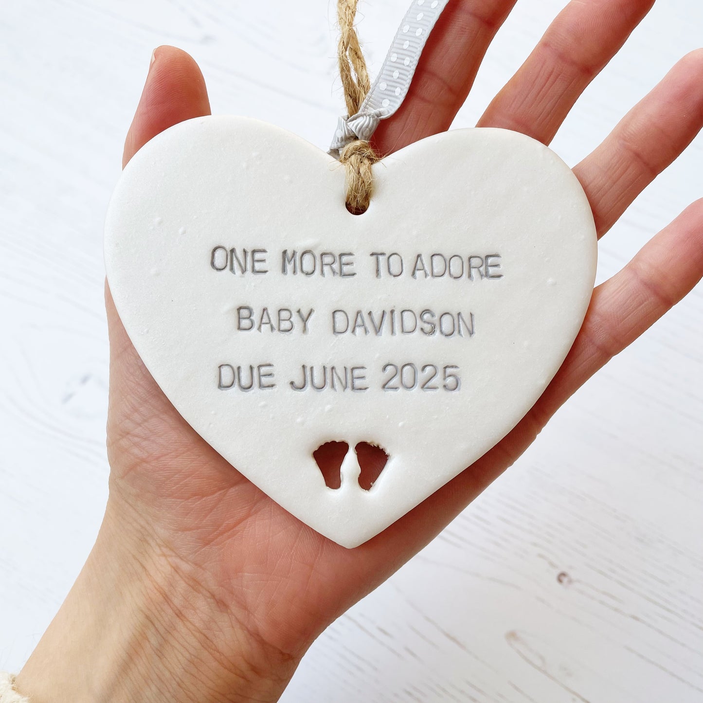 Pearlised white clay hanging heart with baby feet cut out of the bottom and grey personalisation, the heart is personalised with ONE MORE TO ADORE BABY DAVIDSON DUE JUNE 2025