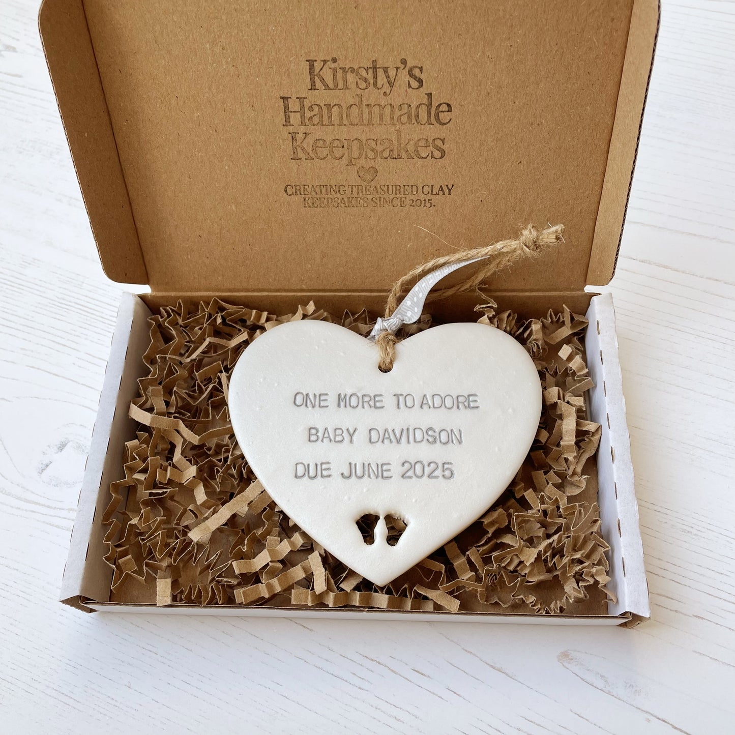 Pearlised white clay hanging heart with baby feet cut out of the bottom and grey personalisation, the heart is personalised with ONE MORE TO ADORE BABY DAVIDSON DUE JUNE 2025