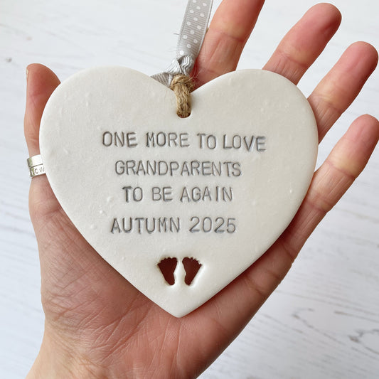 Pearlised white clay hanging heart with baby feet cut out of the bottom and grey personalisation, the heart is personalised with ONE MORE TO LOVE GRANDPARENTS TO BE AGAIN AUTUMN 2025