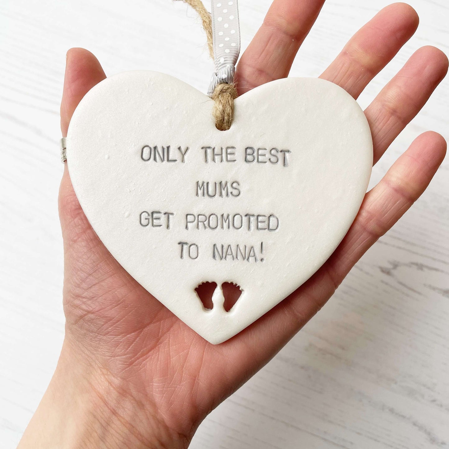 Pearlised white clay hanging heart with baby feet cut out of the bottom and grey personalisation, the heart is personalised with ONLY THE BEST MUMS GET PROMOTED TO NANA!