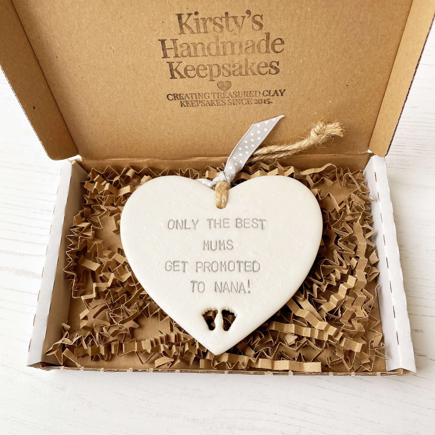 Pearlised white clay hanging heart with baby feet cut out of the bottom and grey personalisation, the heart is personalised with ONLY THE BEST MUMS GET PROMOTED TO NANA!