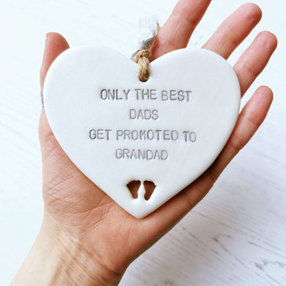 Pearlised white clay hanging heart with baby feet cut out of the bottom and grey personalisation, the heart is personalised with ONLY THE BEST DADS GET PROMOTED TO GRANDAD