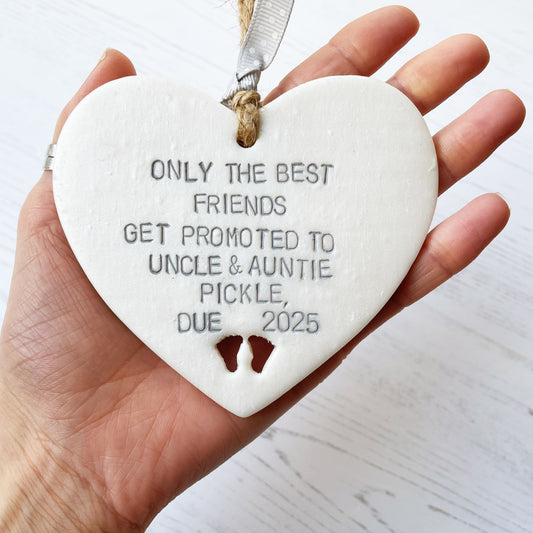 Pearlised white clay hanging heart with baby feet cut out of the bottom and grey personalisation, the heart is personalised with ONLY THE BEST FRIENDS GET PROMOTED TO UNCLE & AUNTIE PICKLE,  DUE 2025