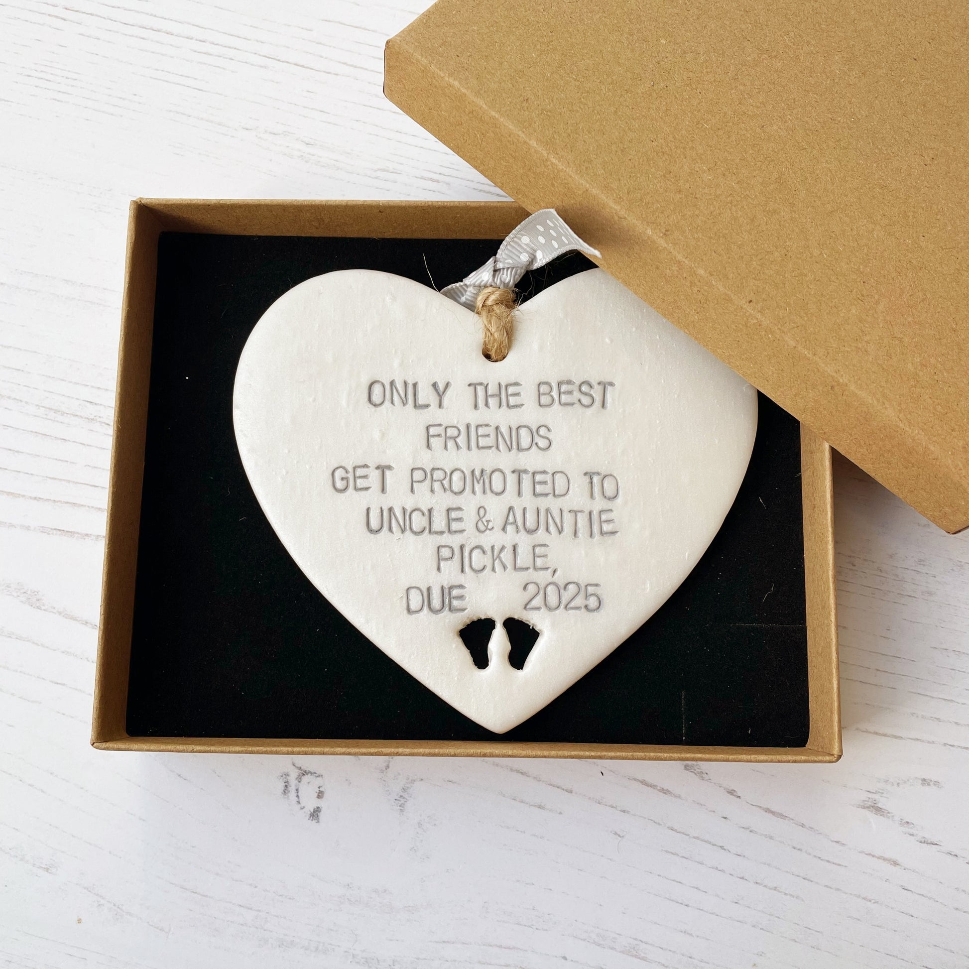 Pearlised white clay hanging heart with baby feet cut out of the bottom and grey personalisation, the heart is personalised with ONLY THE BEST FRIENDS GET PROMOTED TO UNCLE & AUNTIE PICKLE,  DUE 2025