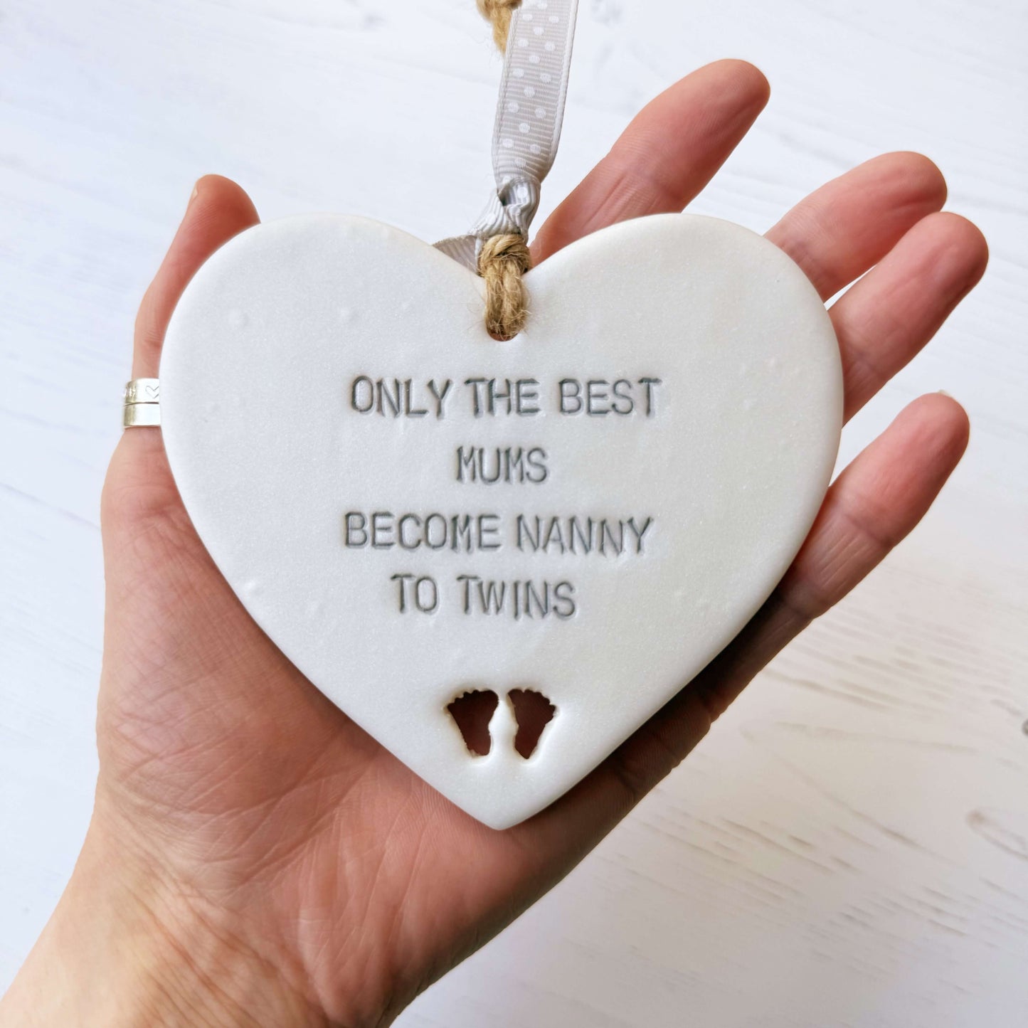 Pearlised white clay hanging heart with baby feet cut out of the bottom and grey personalisation, the heart is personalised with ONLY THE BEST MUMS BECOME NANNY TO TWINS