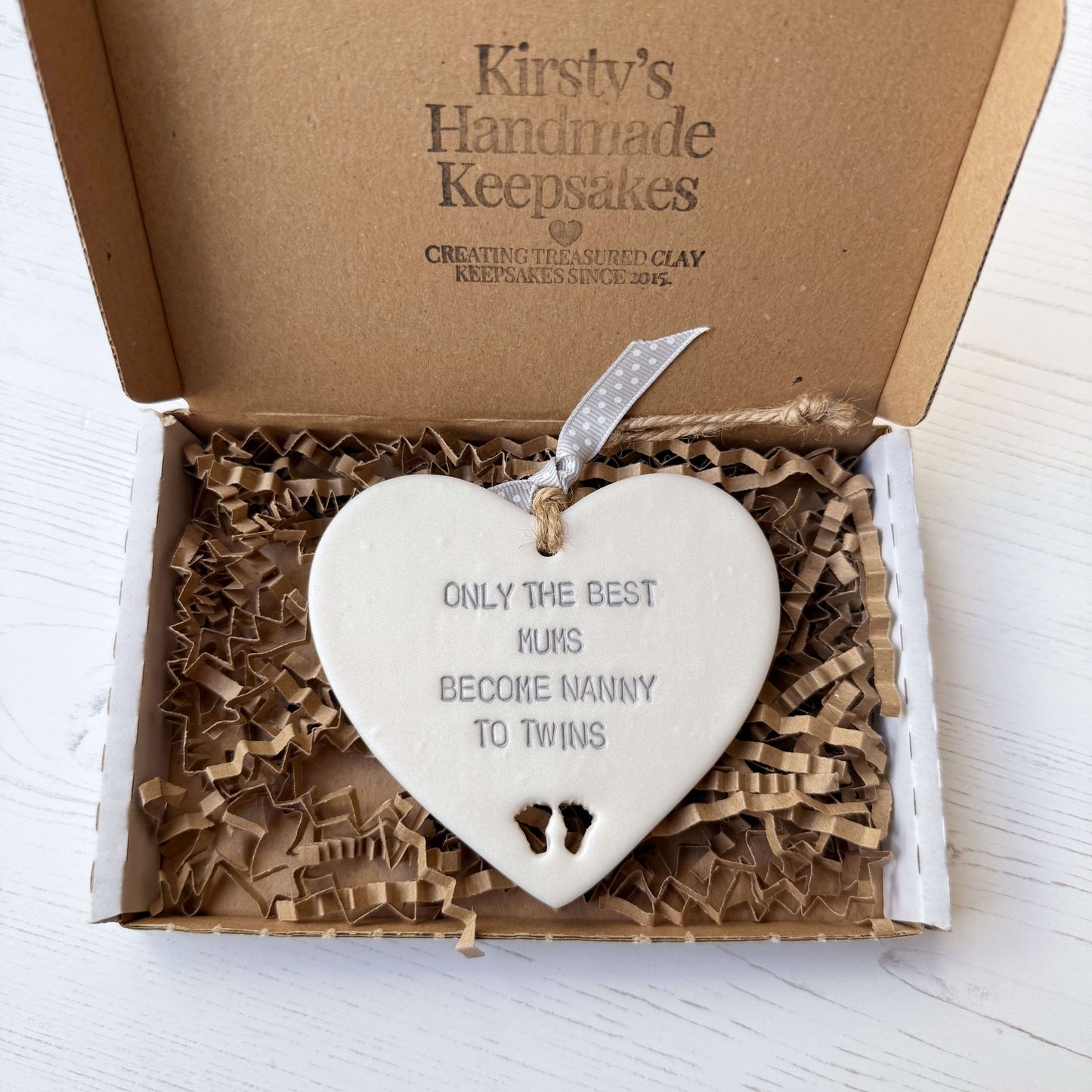 Pearlised white clay hanging heart with baby feet cut out of the bottom and grey personalisation, the heart is personalised with ONLY THE BEST MUMS BECOME NANNY TO TWINS