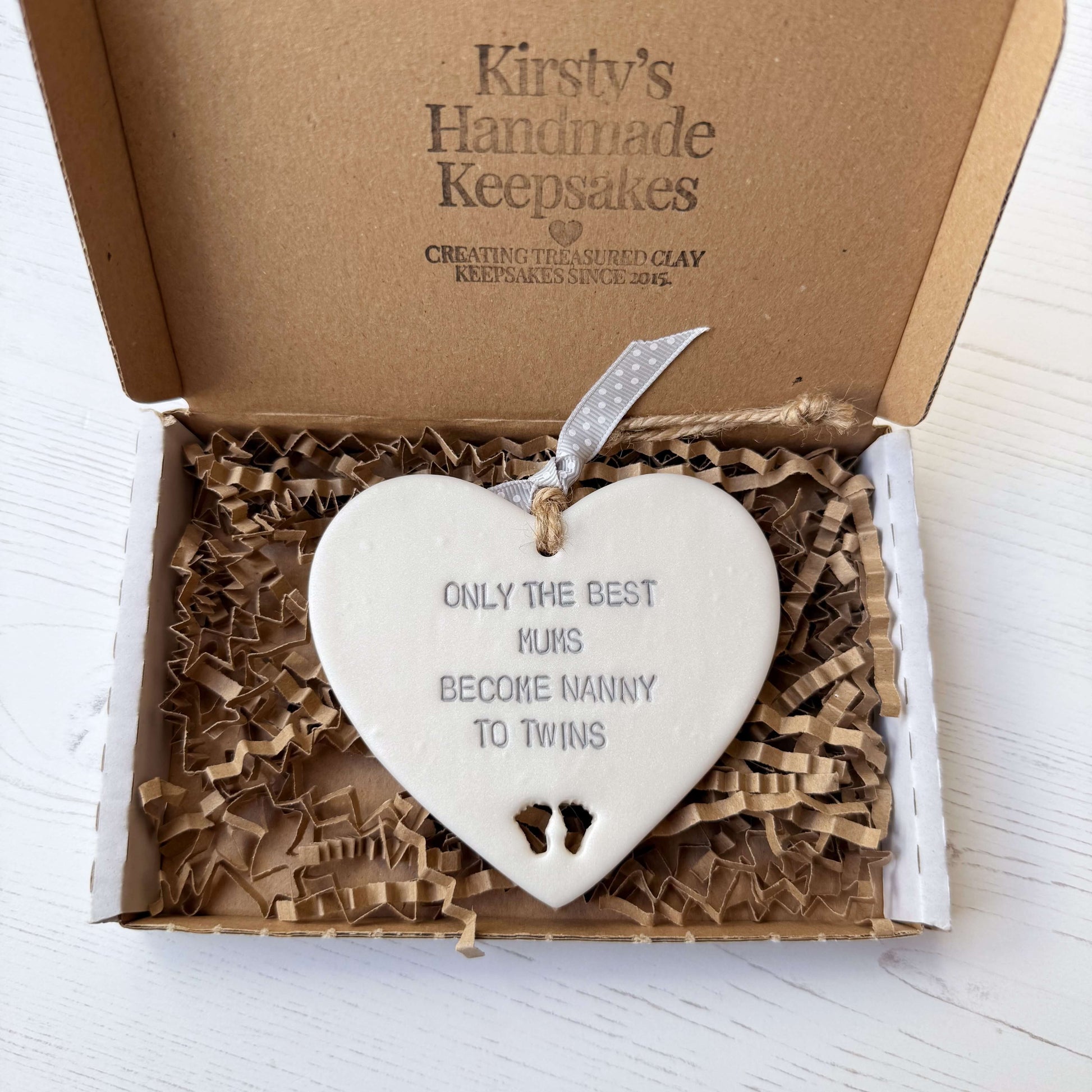 Pearlised white clay hanging heart with baby feet cut out of the bottom and grey personalisation, the heart is personalised with ONLY THE BEST MUMS BECOME NANNY TO TWINS